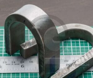 Teste De Dobramento 21 Peça metálica dobrada sobre uma superfície quadriculada, utilizada em testes de resistência mecânica.