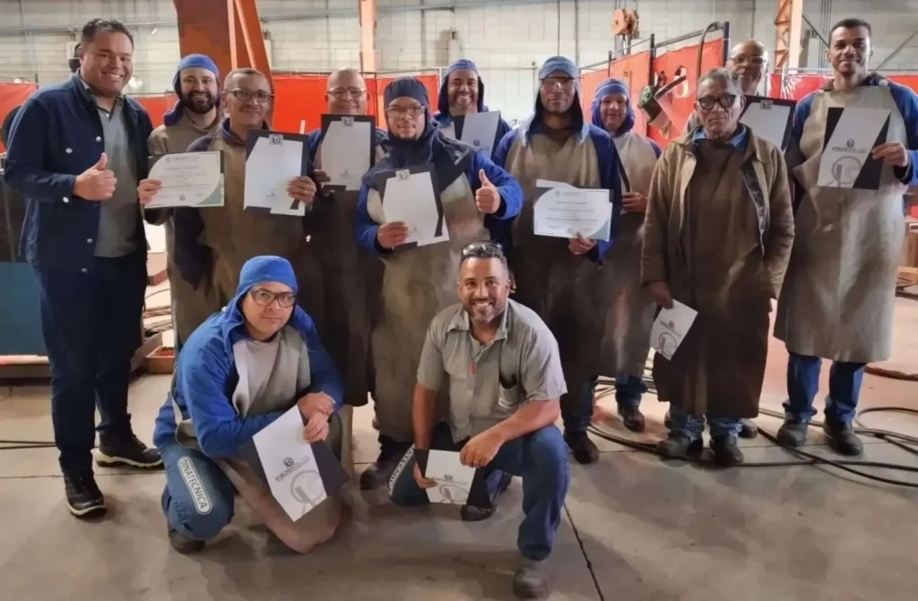 Grupo em ambiente industrial com uniformes e certificados, celebrando treinamento concluído com instrutor.