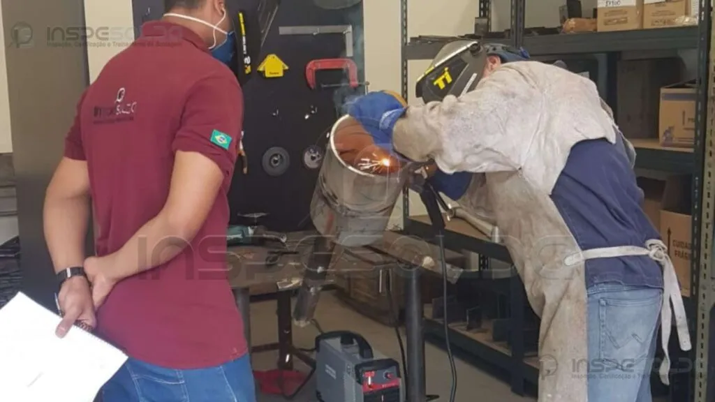 home-qualificacao-soldagem-acompanhamento Técnico observa e analisa soldador em trabalho com equipamento cilíndrico em bancada industrial.